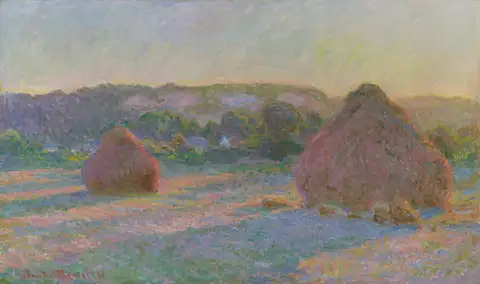Two large stacks of wheat in a field at the end of a summer day, with hills and houses behind.